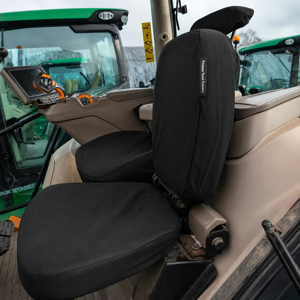 Black seat covers fitted over a tractor cabin seat, shown installed in a real vehicle interior with other tractors visible in the background.