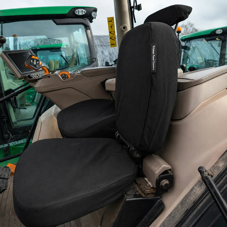 Black seat covers fitted over a tractor cabin seat, shown installed in a real vehicle interior with other tractors visible in the background.